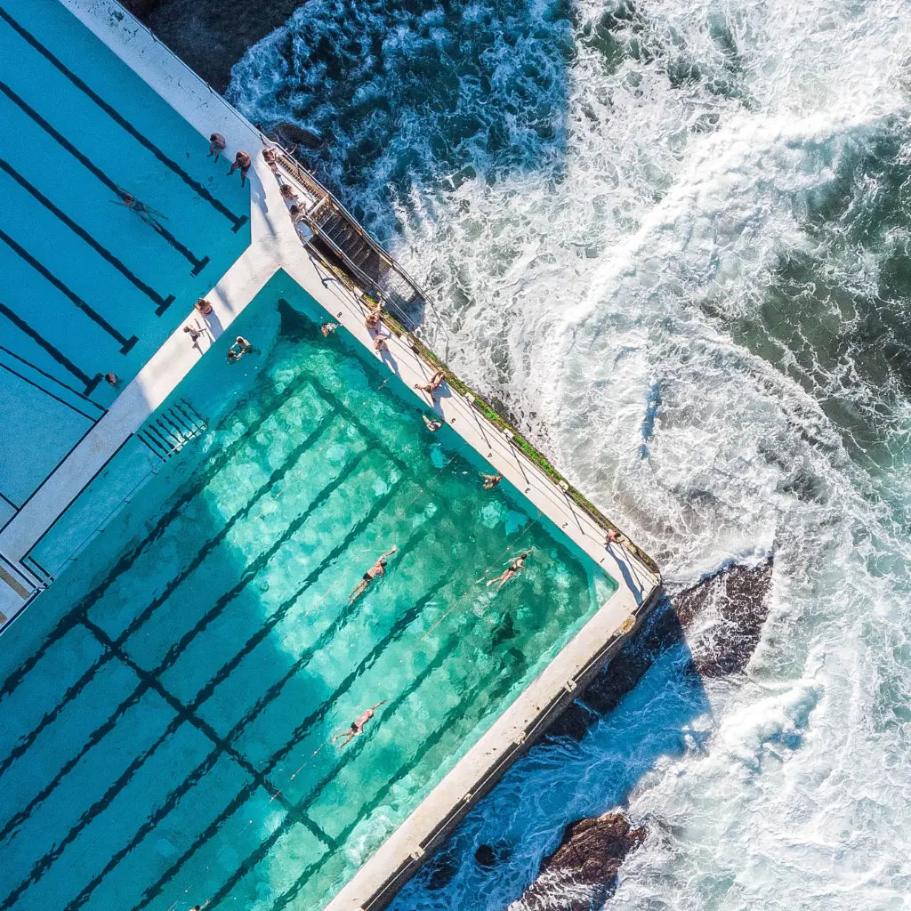 Aerial view of a Bondi Icebergs swimming pool adjacent to the ocean, with swimmers in the turquoise pool and waves crashing around the concrete edge.