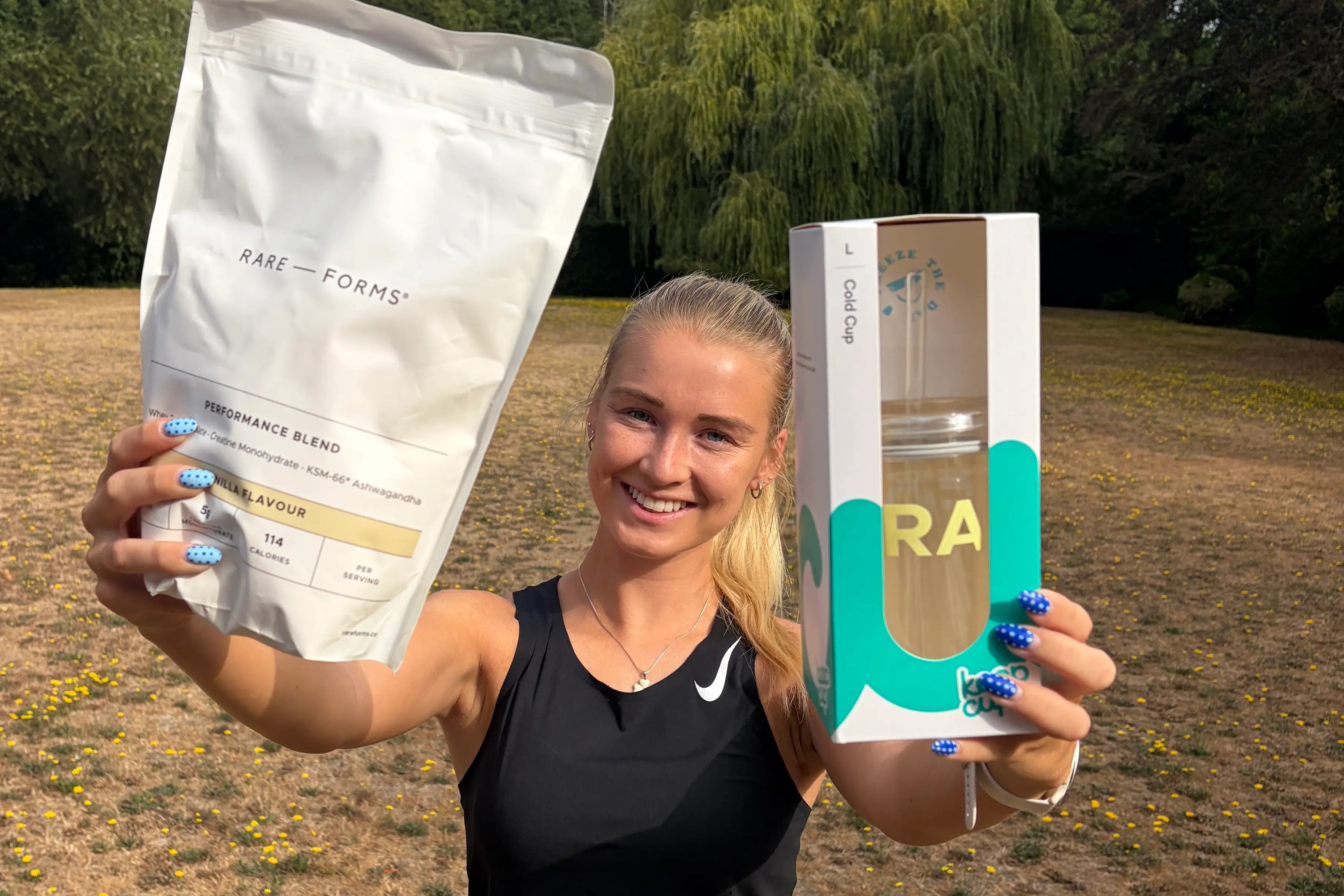 A woman outdoors in a grassy field holds up a large white bag of Rase Forms protein powder and a boxed bottle of water. Trees and blue sky are visible in the background.